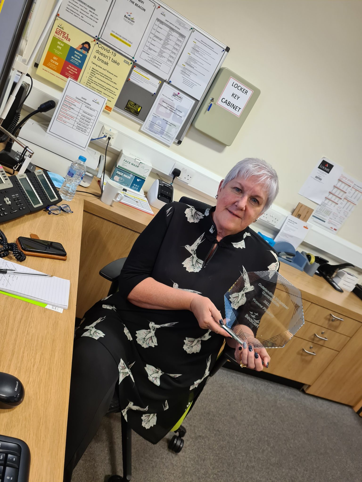 This shows Yvonne sat at her desk in Beacon's reception area. She has short hair and is dressed all in black with a tunic style top that has large grey flowers. She's holding her award, made of glass in a hexagon shape and is smiling towards the camera. 